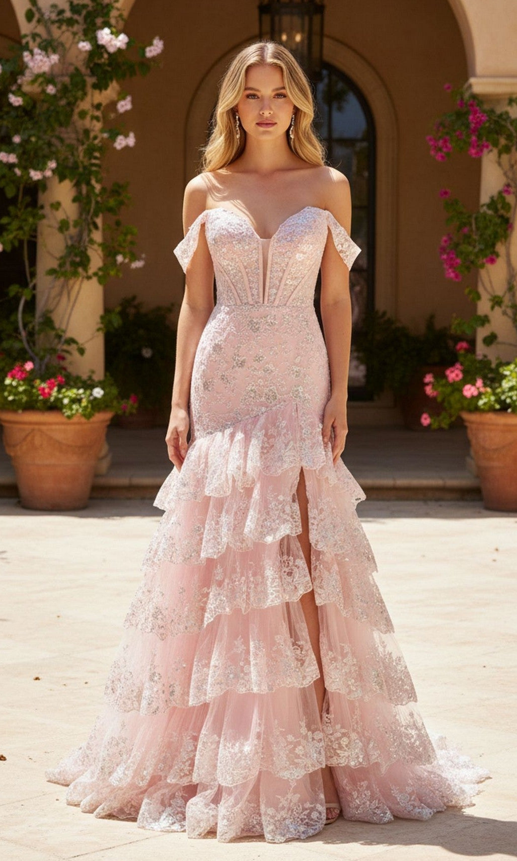 A woman wearing the Long Prom Dress: PromGirl Exclusive 250068 in blush stands outdoors in front of a building decorated with potted plants and flowers.